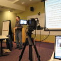 Brendan speaking in front of a projector screen, videocamera in the foreground
