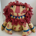 A stuffed toy lion made of yellow cotton printed with red flowers. He has a big grin with blue teeth, swirly red cheeks, and a mane made of loops of sewn cotton strips
