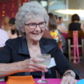 An older lady with curly gray hair and glasses grins as she holds a glass with a sugared rim, sitting at a colorful table
