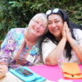 A Canadian woman with short white hair and an Indian woman with long black hair sitting at a table, leaning their heads together and smiling like the old friends they are