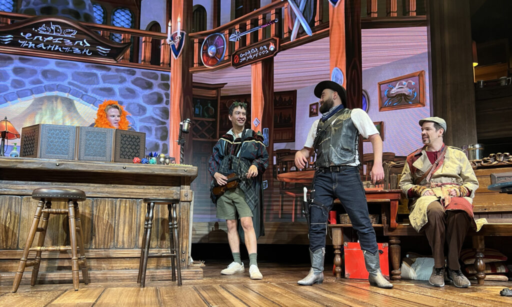 front-row view of the stage at Twenty Sided Tavern. Center stage is a bar with barstools, behind it stands the Dungeon Master directing the campaign and playing bit parts, at the moment he has an orange feather boa wrapped around his head. Three other actors improvise a scene based on the characters the audience selected for them.