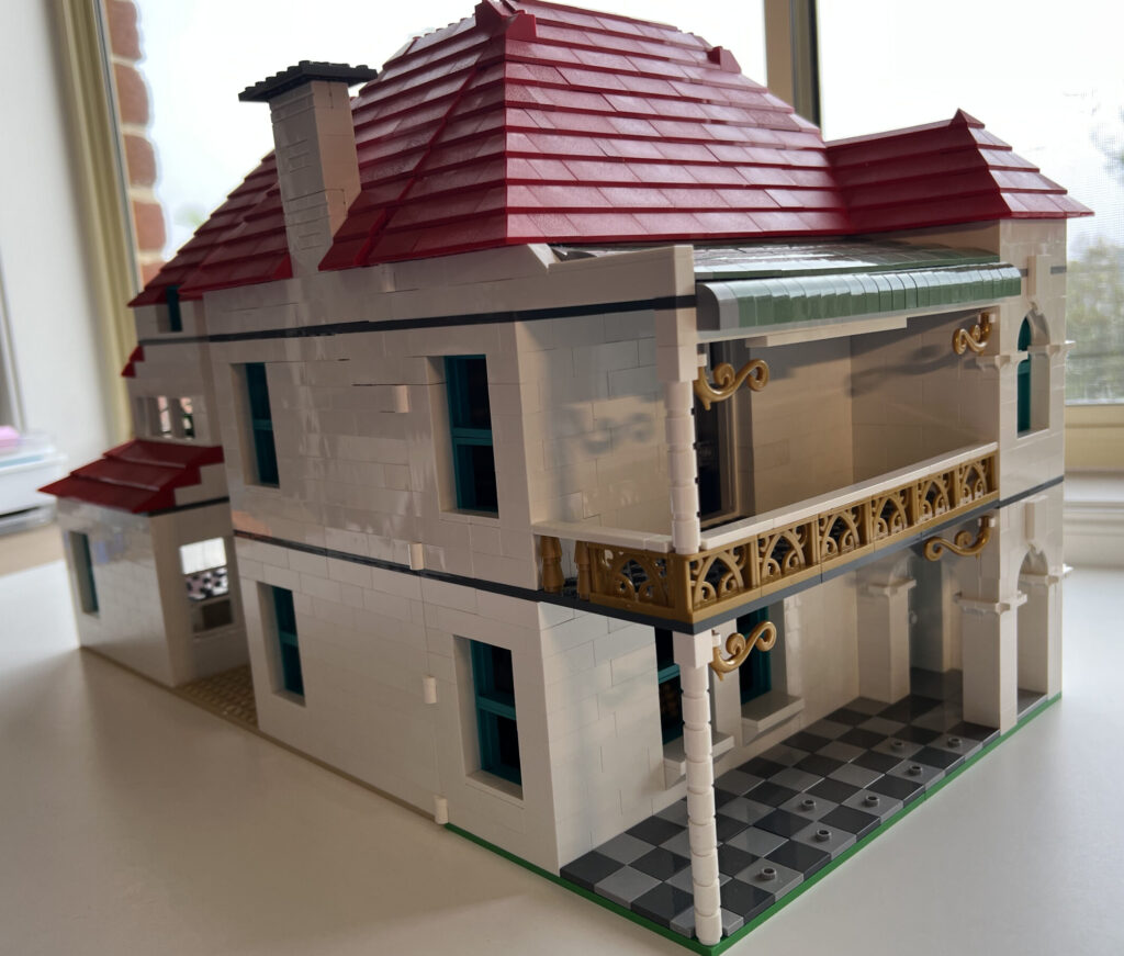 a white Lego house with a steep red room, a dull green roof over the front balcony, and a tower-like bit on the front with its own peaked roof
